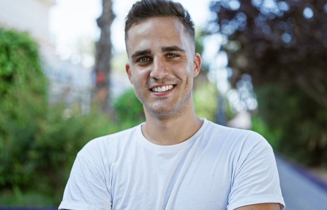 A man wearing a white t-shirt stands outdoors, smiling at the camera, with greenery and trees in the blurred background.
