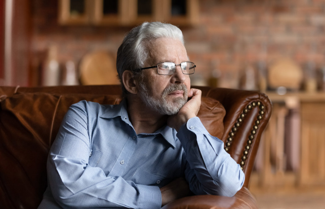 Older man with gray hair and glasses sitting on a leather sofa, gazing thoughtfully into the distance, wearing a light blue shirt. Brick wall and wooden shelves in the blurred background.